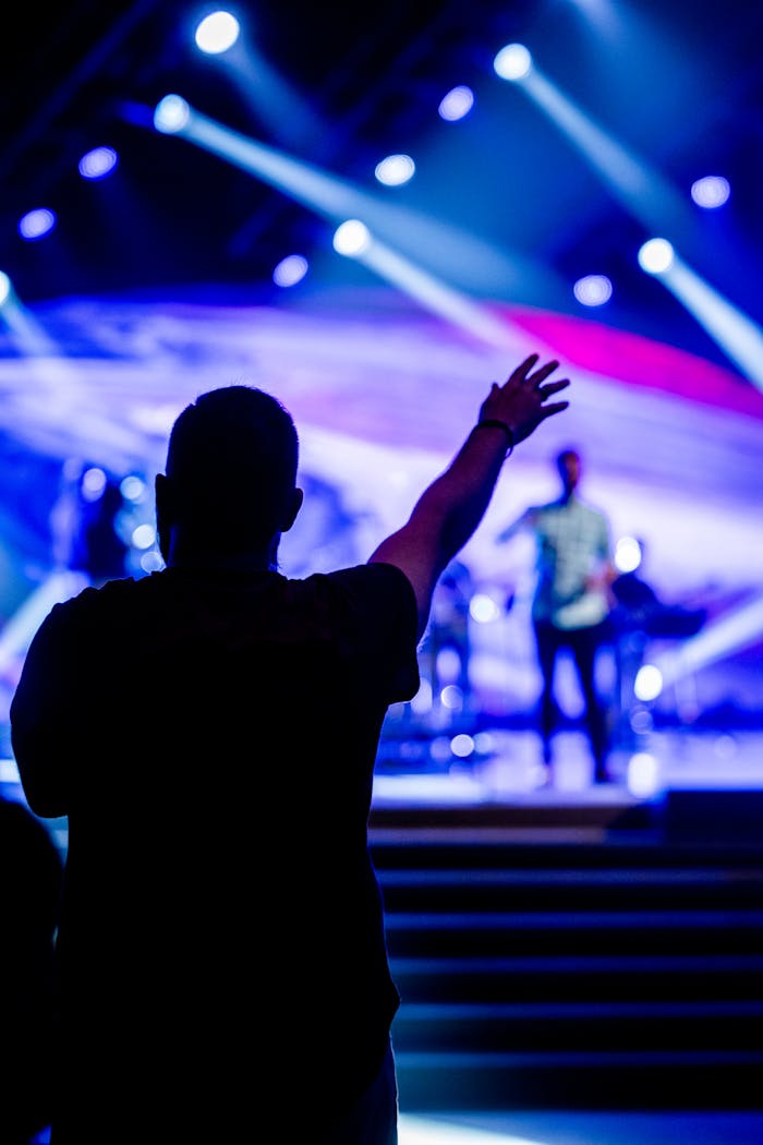 About Silhouette of a man raising his hand at an energetic live concert indoors with vibrant stage lighting.